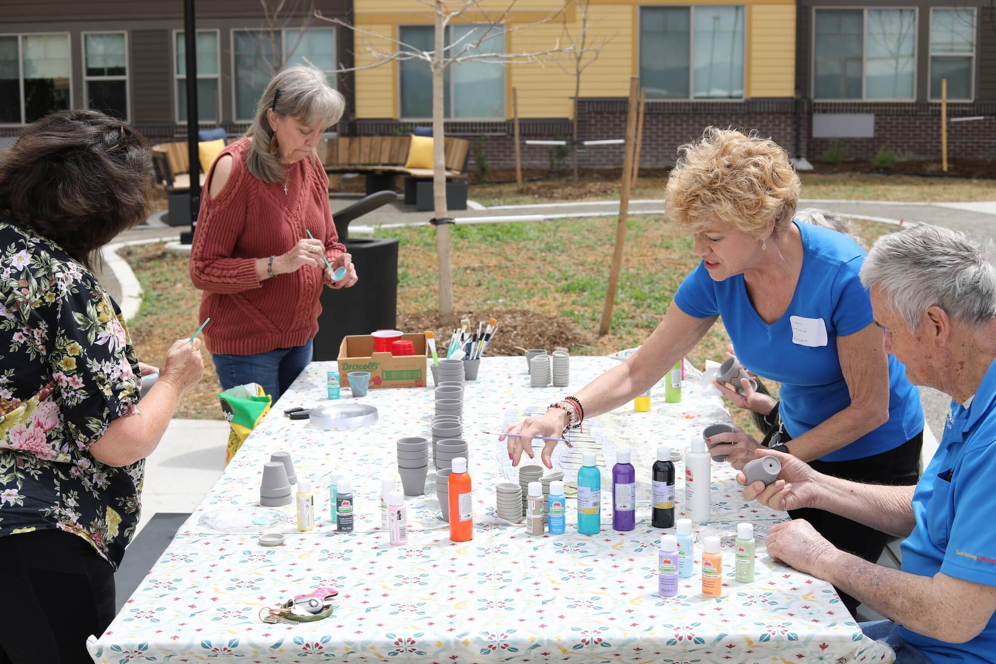 People painting small pots at an outdoor table covered with art supplies, including various paint bottles and brushes