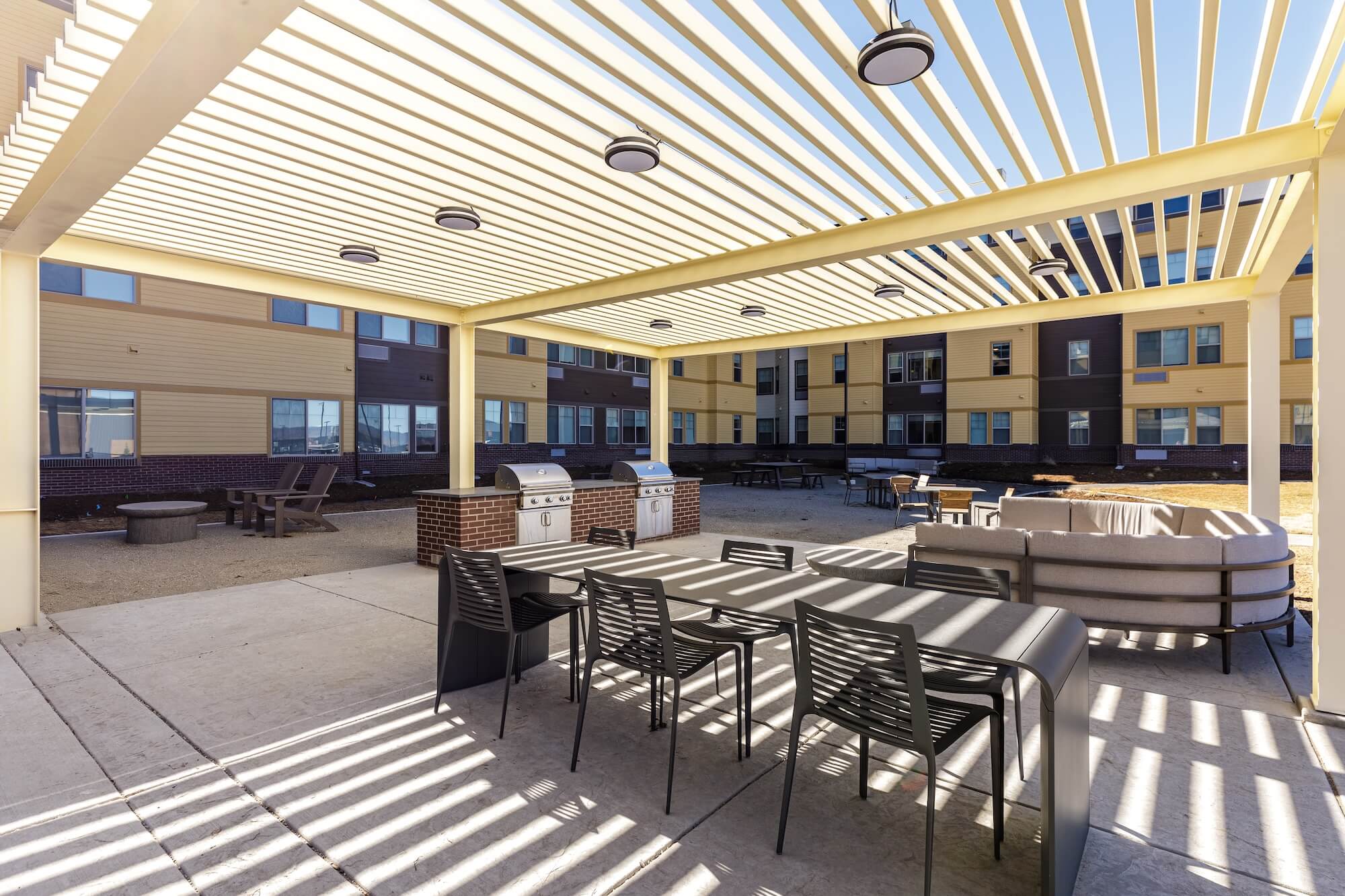 Outdoor patio with pergola, dining tables, chairs, grills, and seating area. Apartment building facade in the background