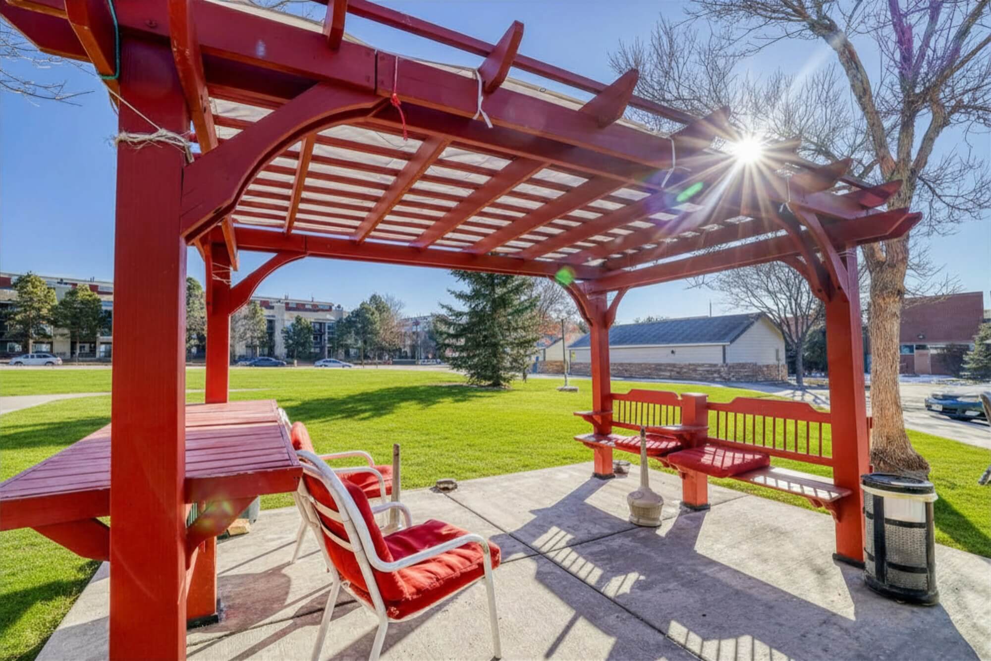 Red wooden gazebo with benches and chairs on a patio in a sunny park setting, surrounded by grass and trees