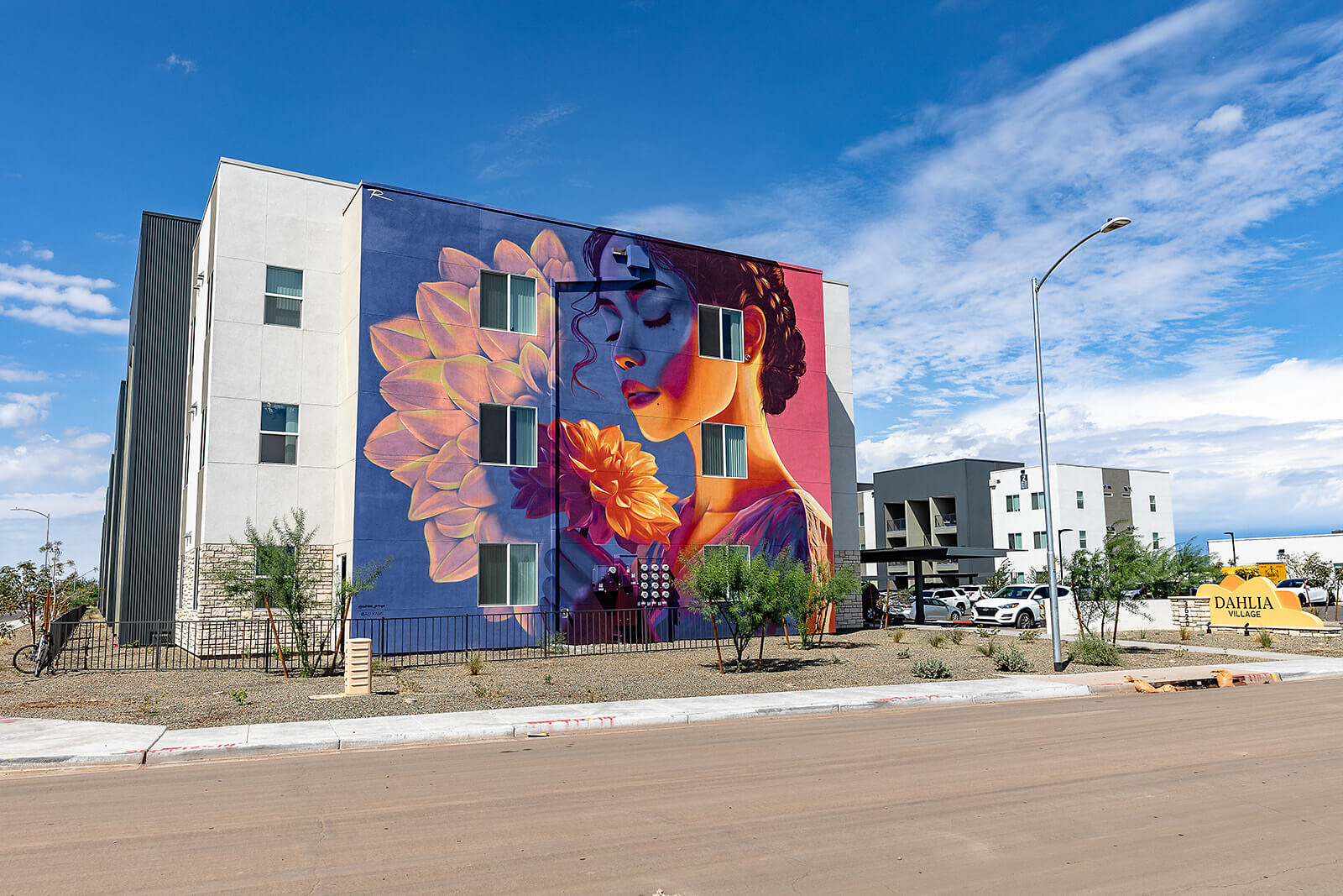 Colorful mural of a woman's face with flowers on a building. Sign reads "DAHLIA VILLAGE" in front of modern apartments