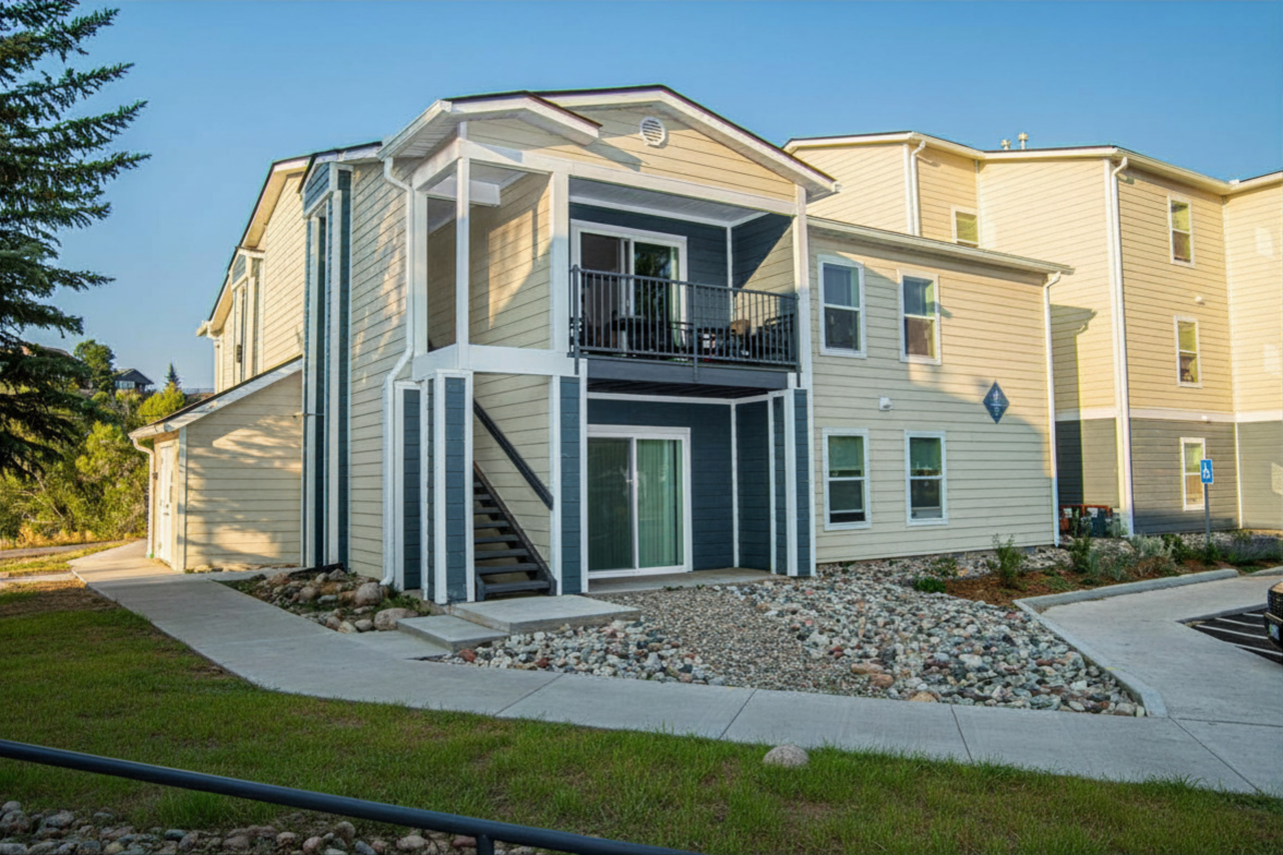 Two-story beige and blue apartment building with a covered balcony, surrounded by a landscaped area with rocks and grass