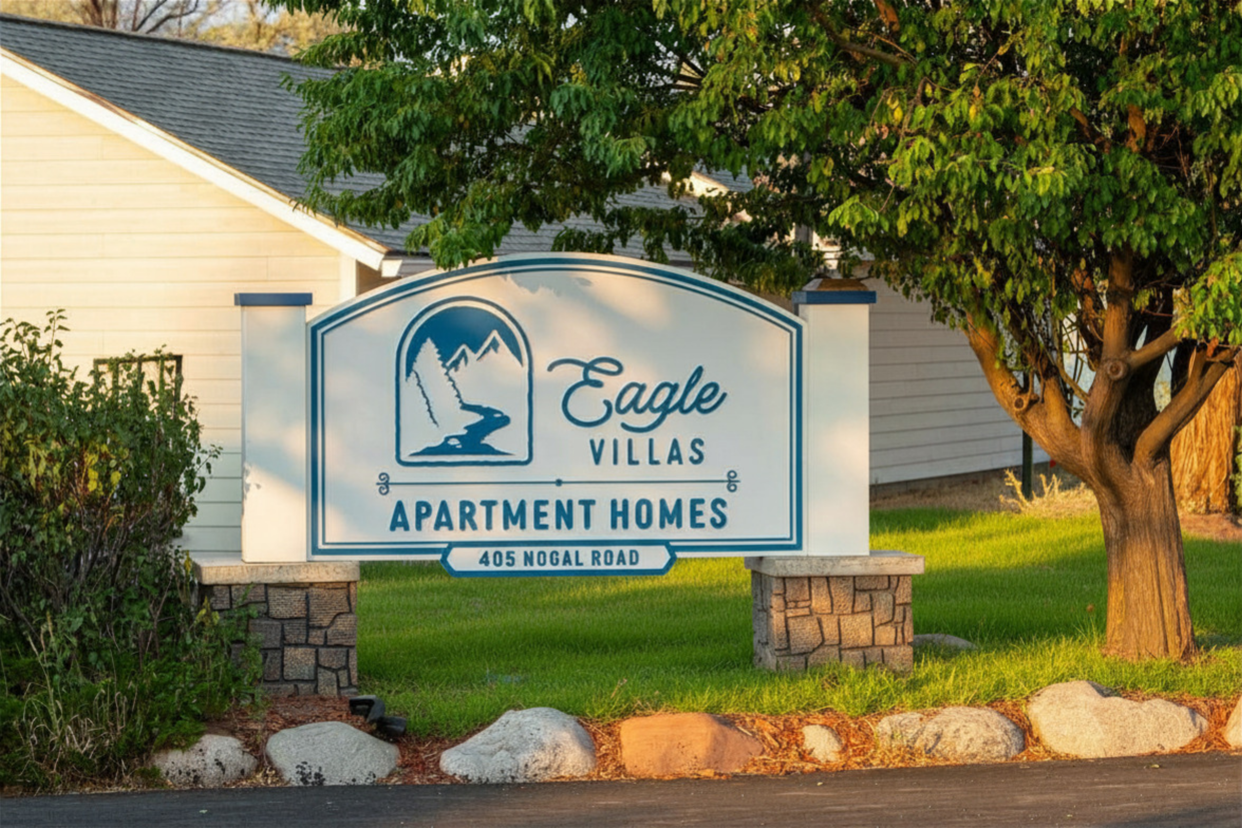 Eagle Villas Apartment Homes sign with a mountain and river graphic, located at 405 Nogal Road, surrounded by greenery