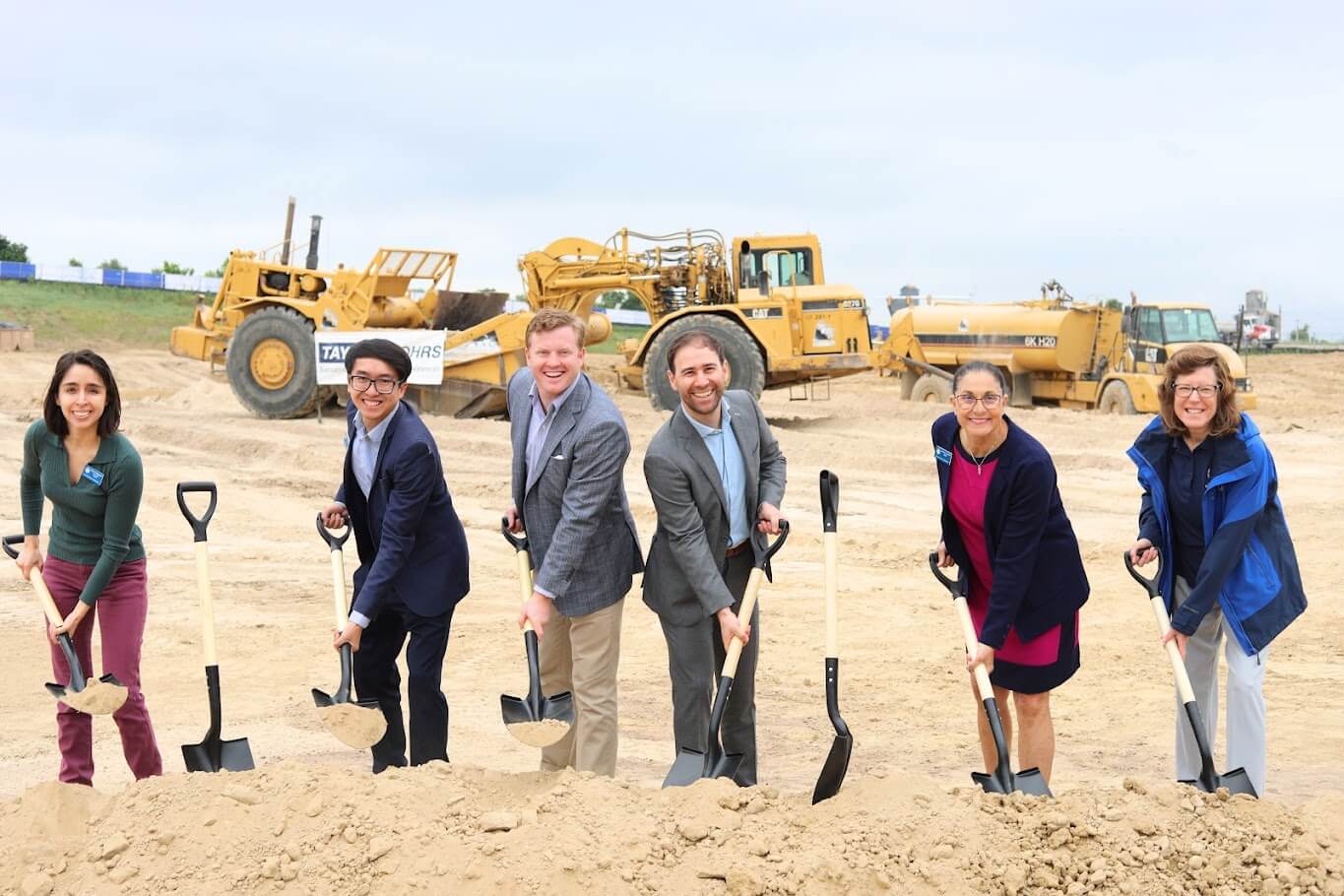 Six people with shovels at a construction site groundbreaking ceremony, with heavy machinery in the background