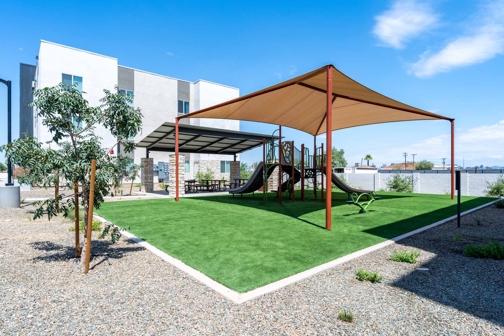 Playground with slides and shade canopy on artificial grass, next to picnic tables under a metal awning, near a building