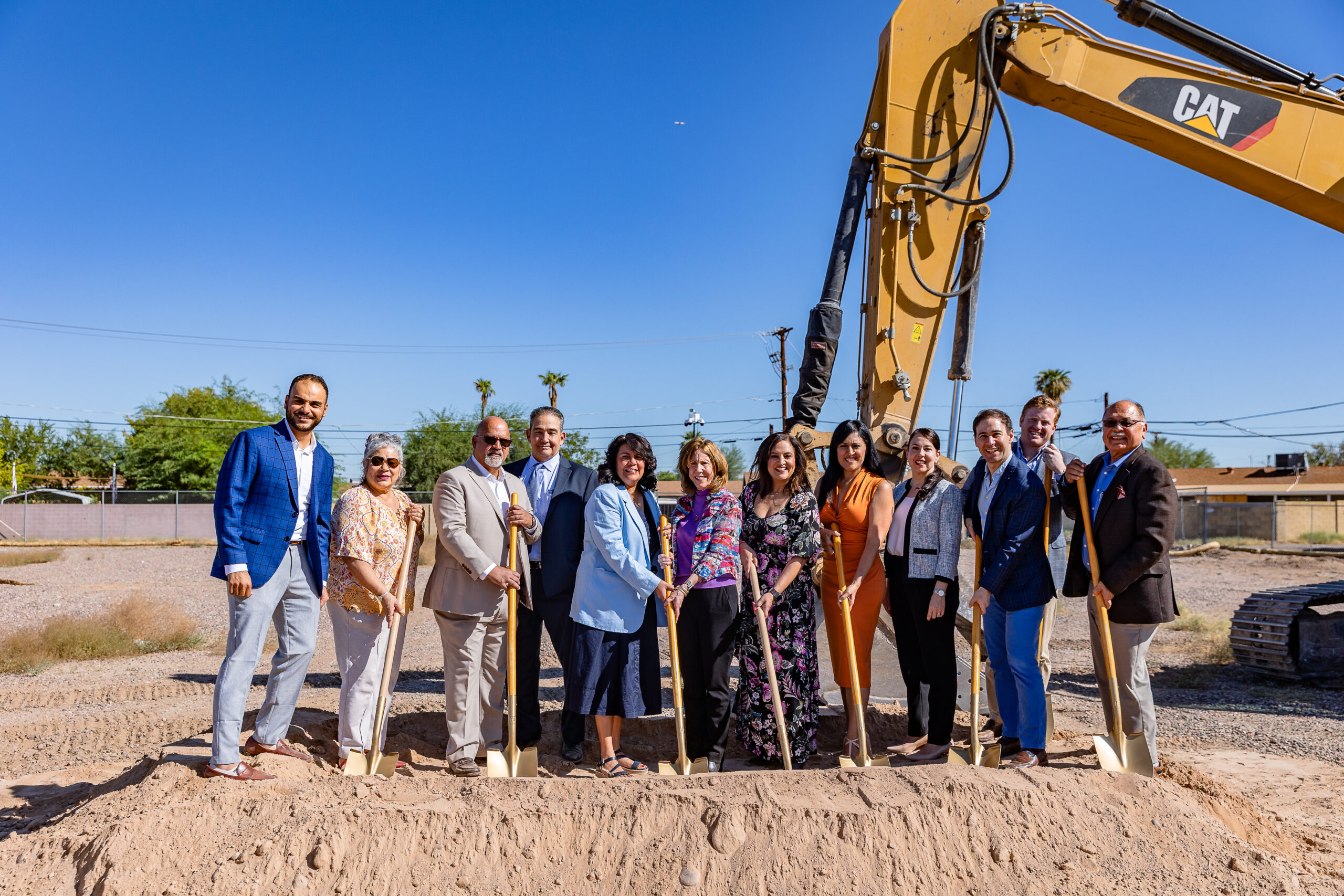 Group of people holding shovels at a groundbreaking ceremony in front of a construction excavator under a clear blue sky