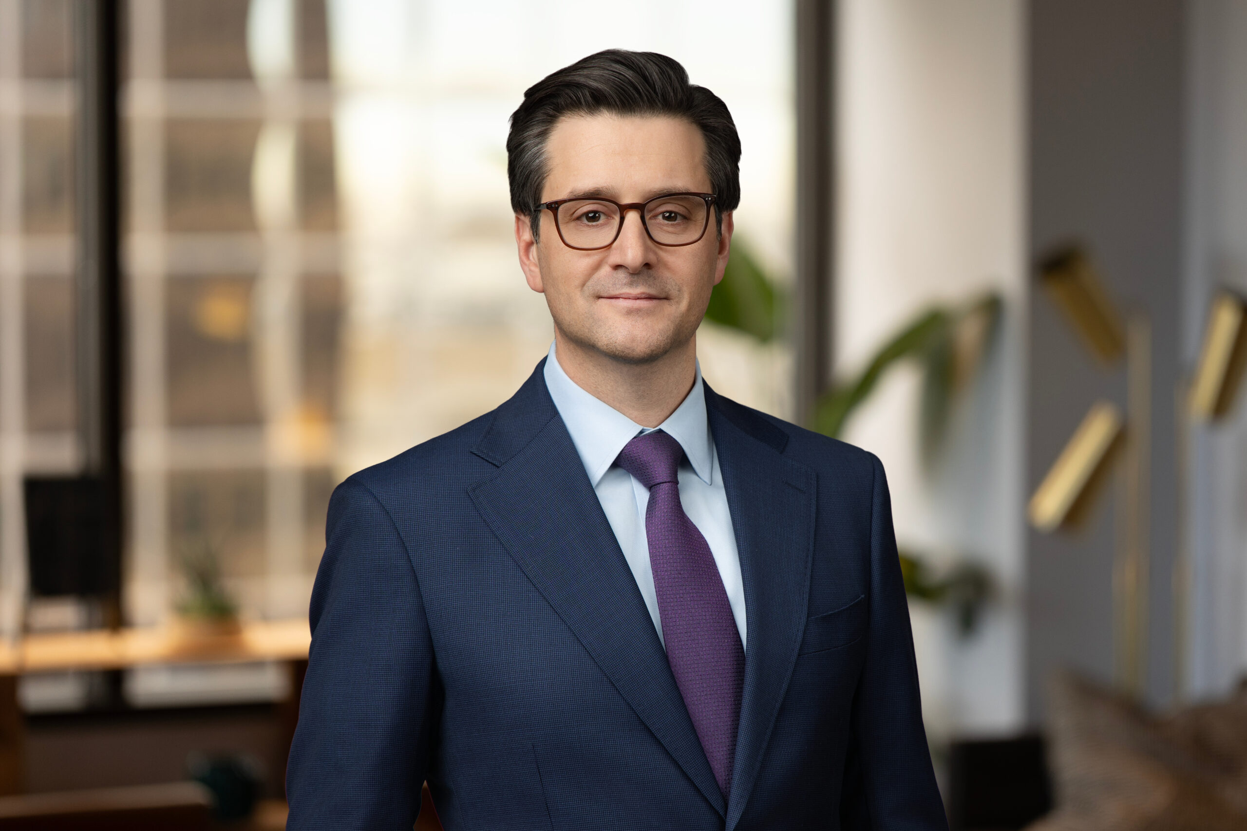 Man in a dark suit and glasses standing in a modern office with large windows and plants in the background