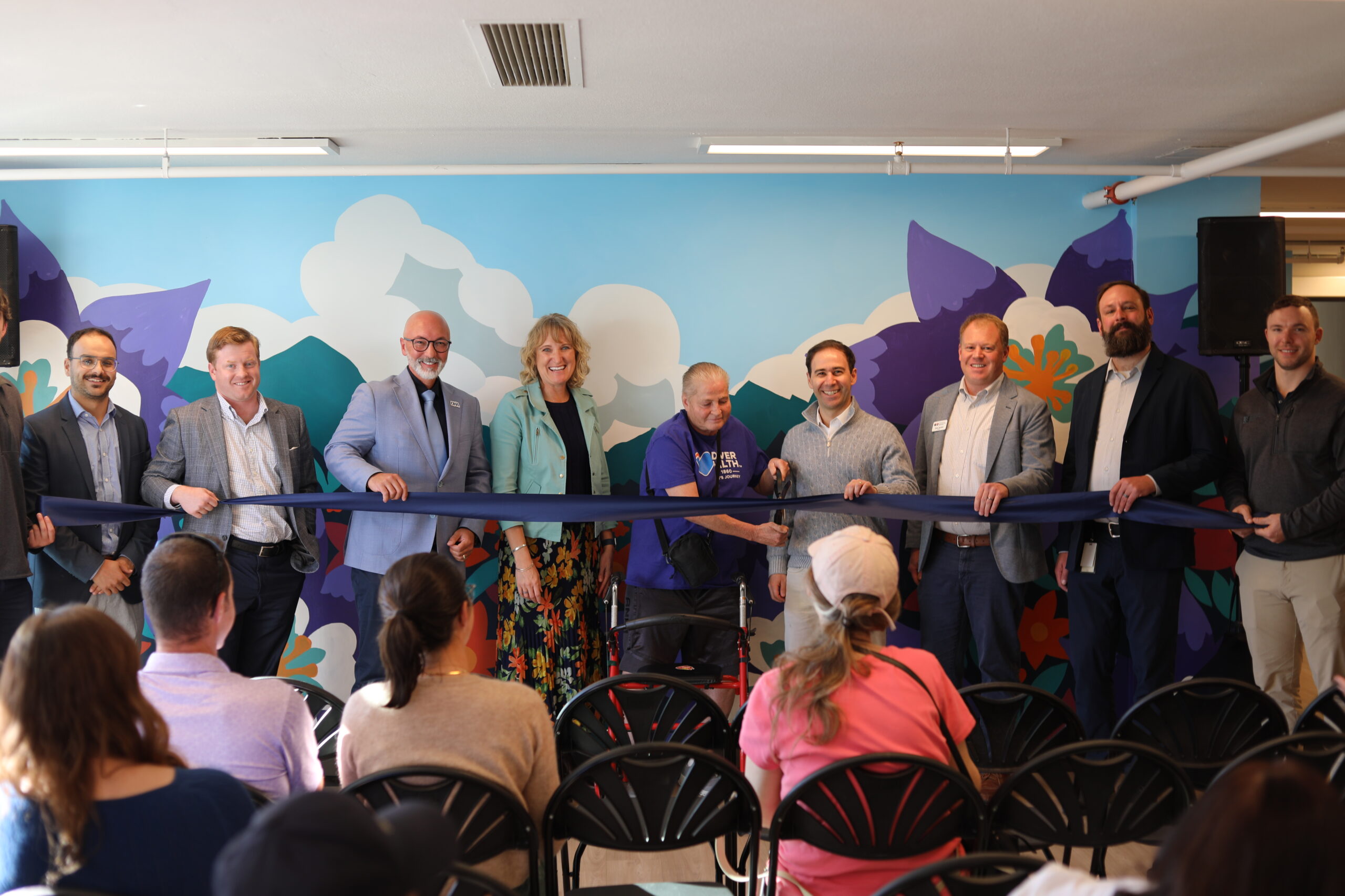 Group of people holding a large blue ribbon at a ceremony, smiling, in front of a colorful mural, with seated audience