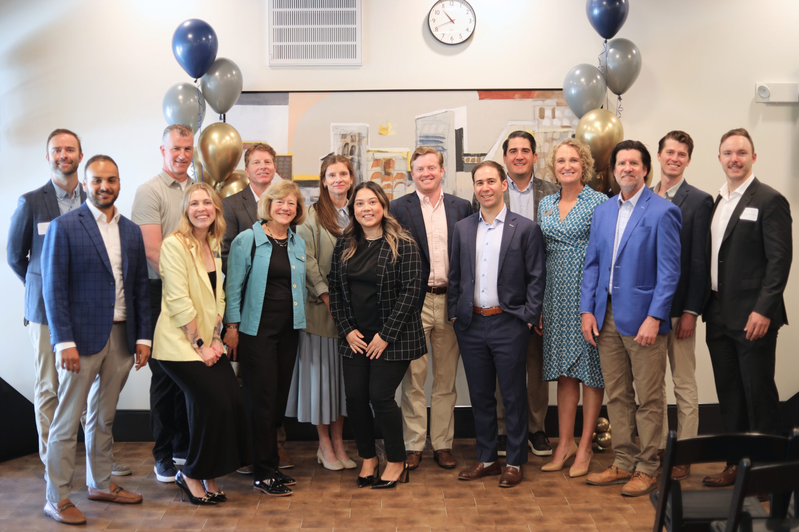 Group of 16 formally dressed people smiling in a room with decorative balloons, a clock, and framed artwork on the wall