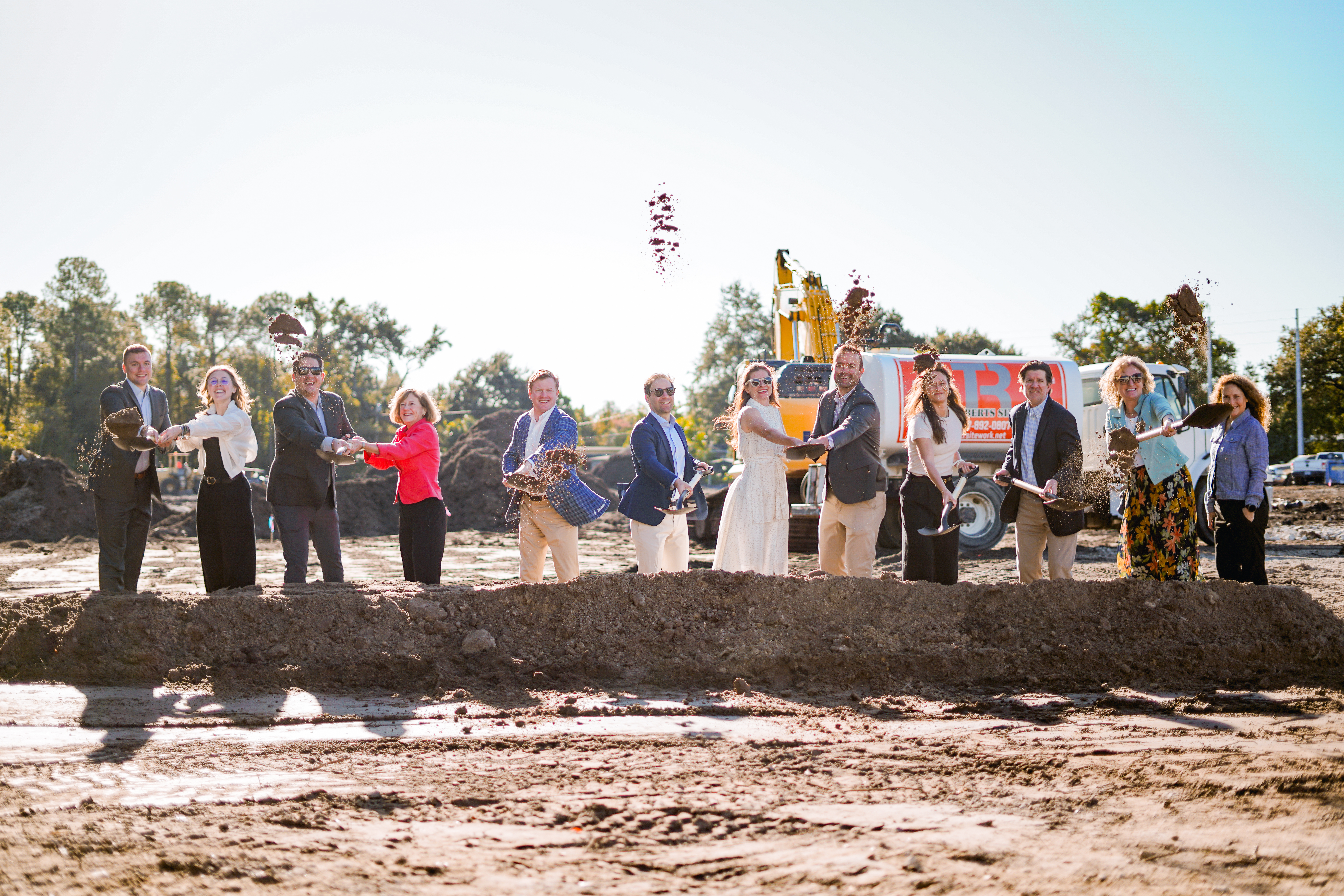 Group at a groundbreaking ceremony, lifting soil with shovels; construction equipment and trees in the background