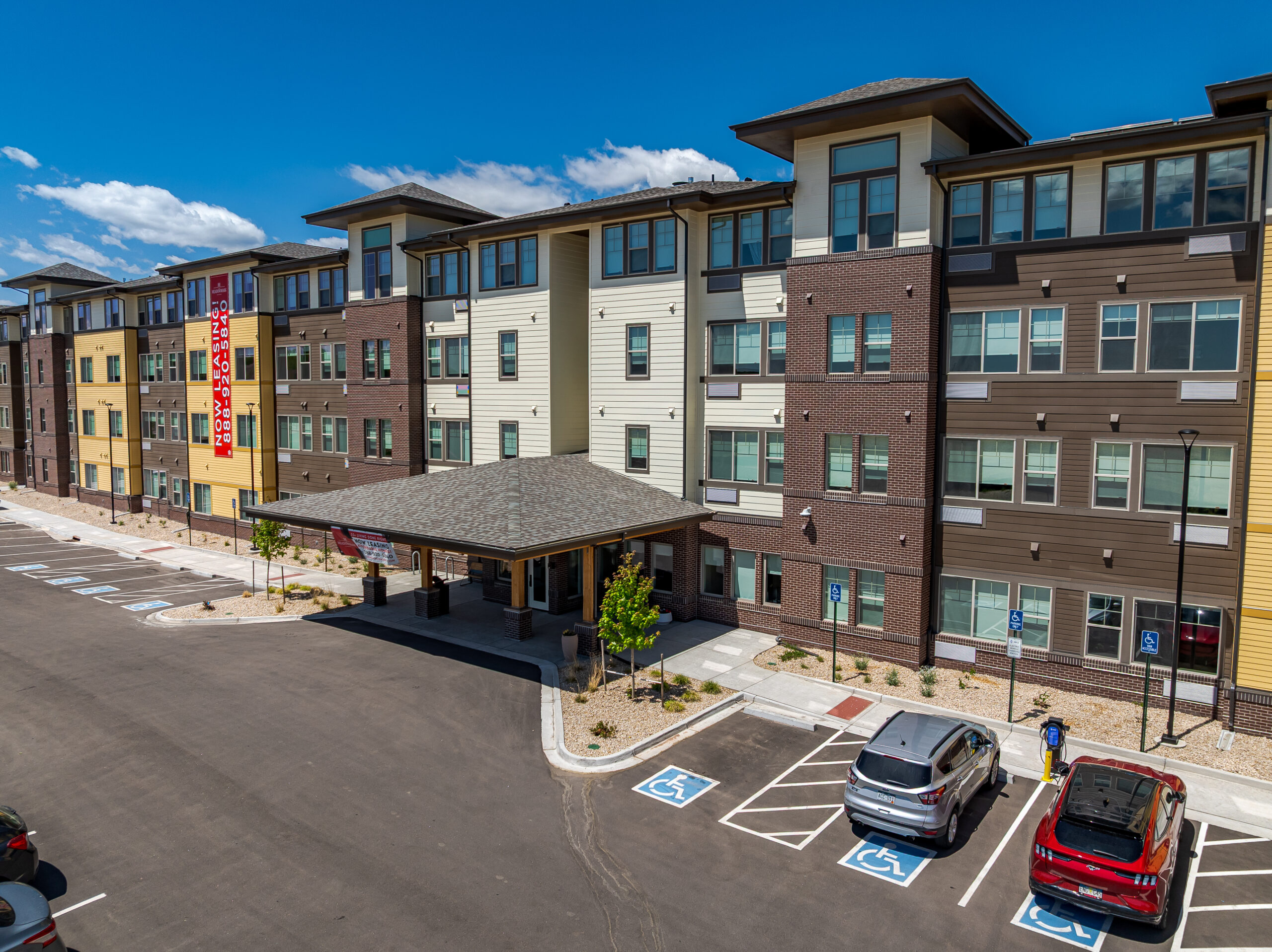 Four-story apartment building with modern design, two cars parked in accessible spaces, and a banner saying "Now Leasing"