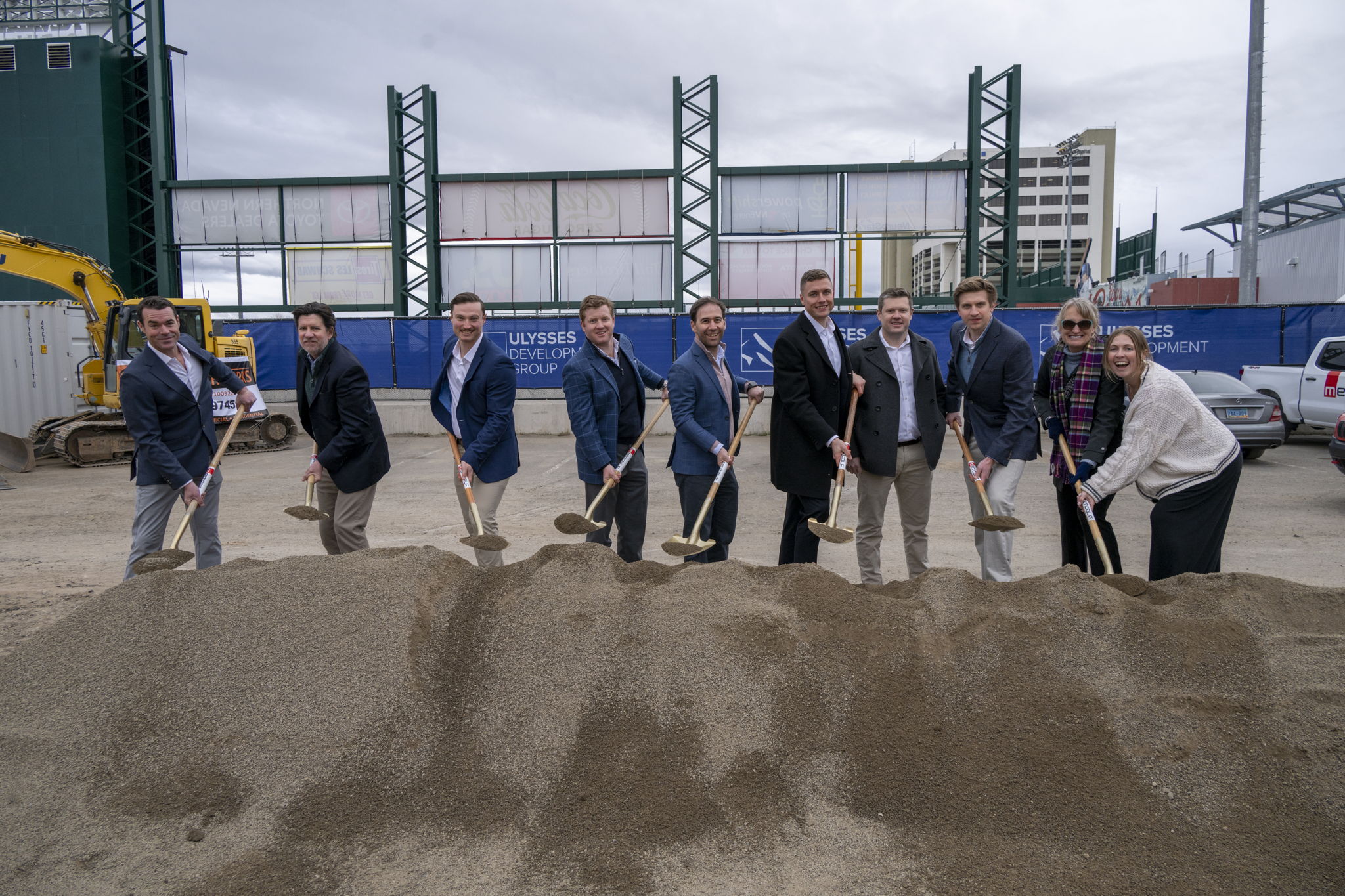 A group of people in formal attire participate in a ceremonial groundbreaking with shovels on a construction site