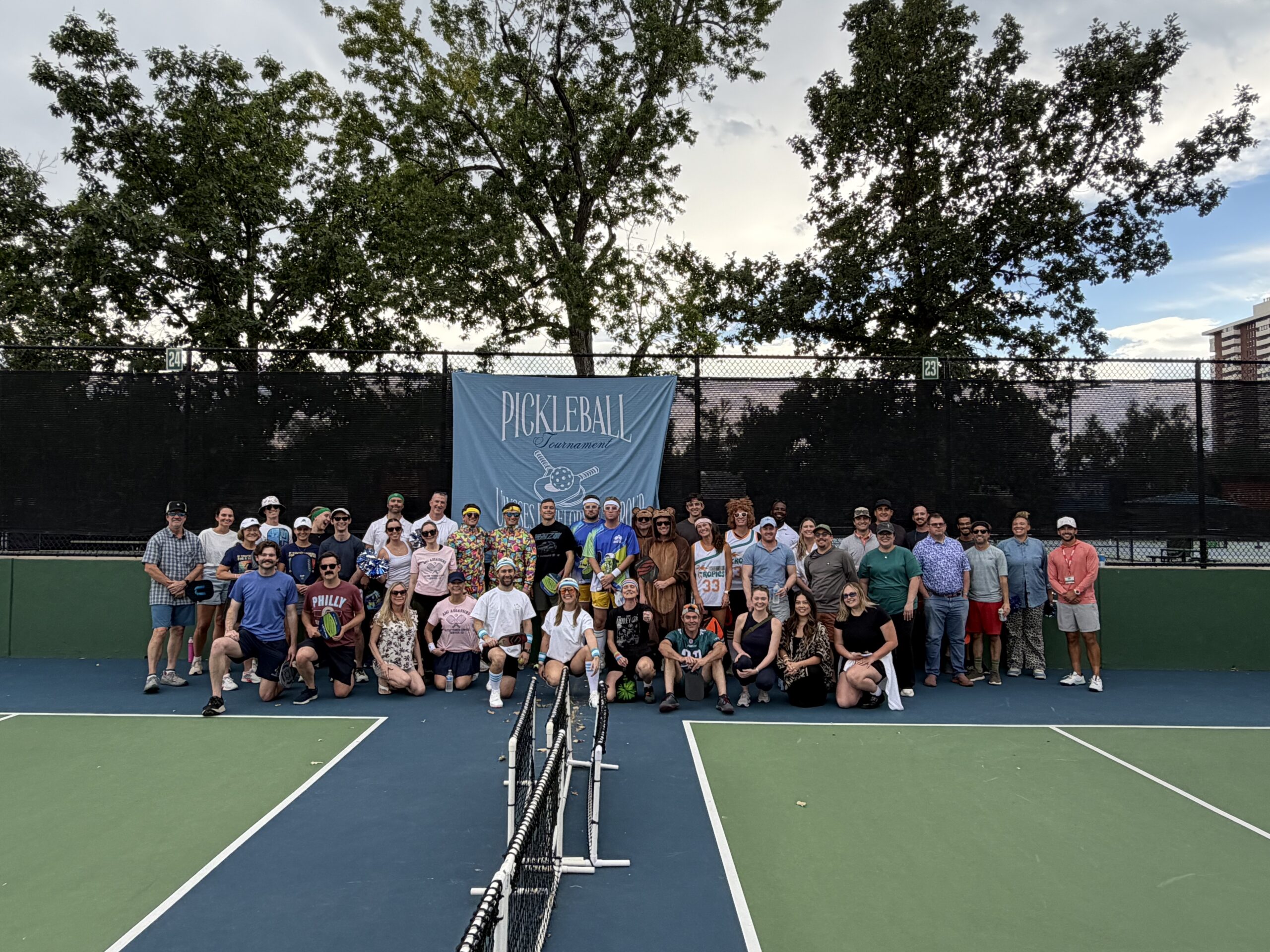 Group of people on a tennis court posing in front of a banner that reads "Pickleball Tournament" with trees in the background