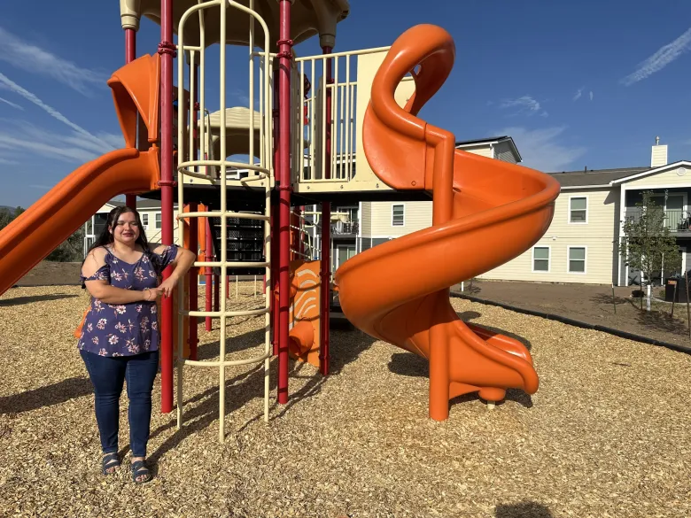 Smiling woman stands by a large playground slide on a sunny day, with residential buildings in the background