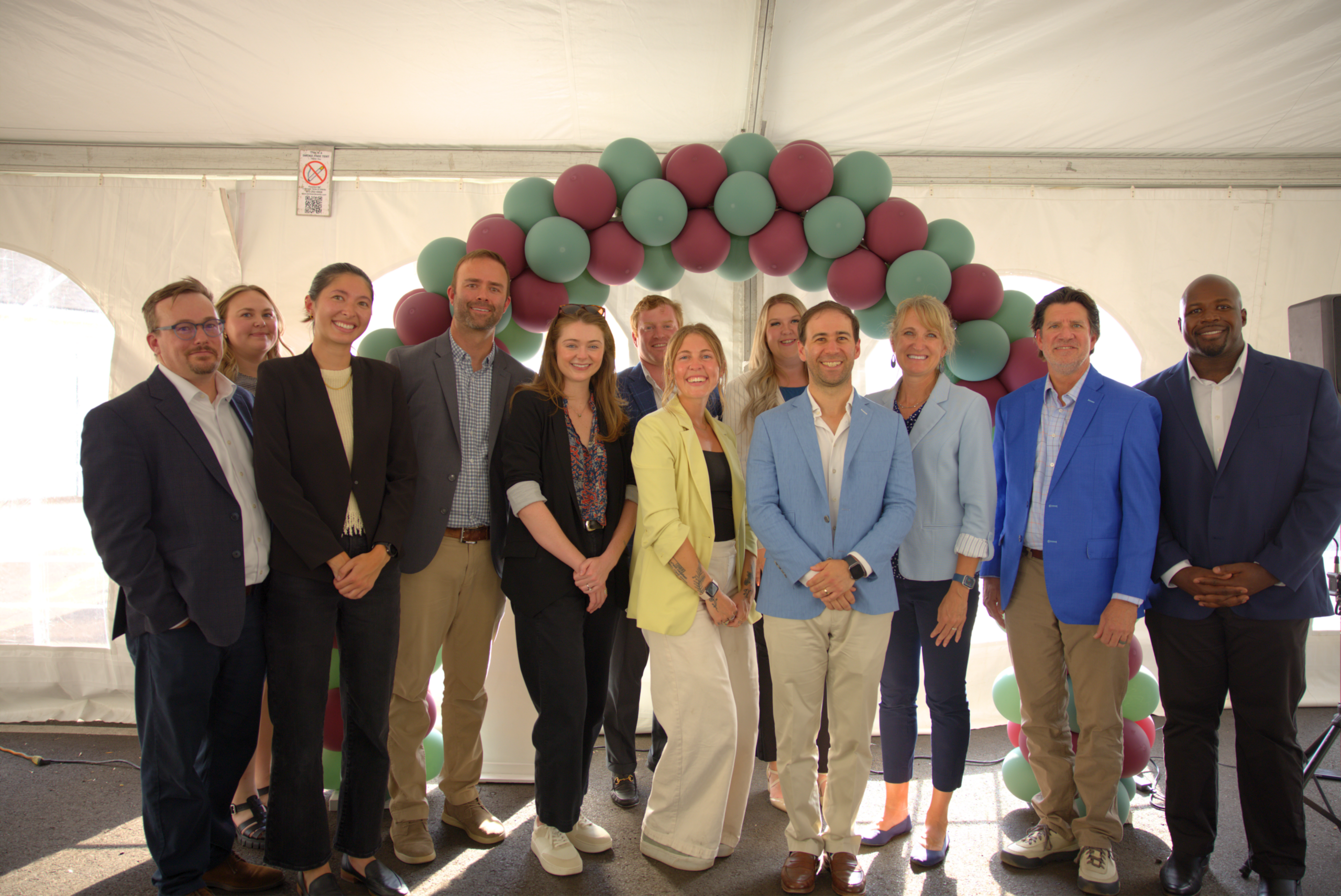 Group of people smiling under a balloon arch inside a tent, wearing business-casual attire