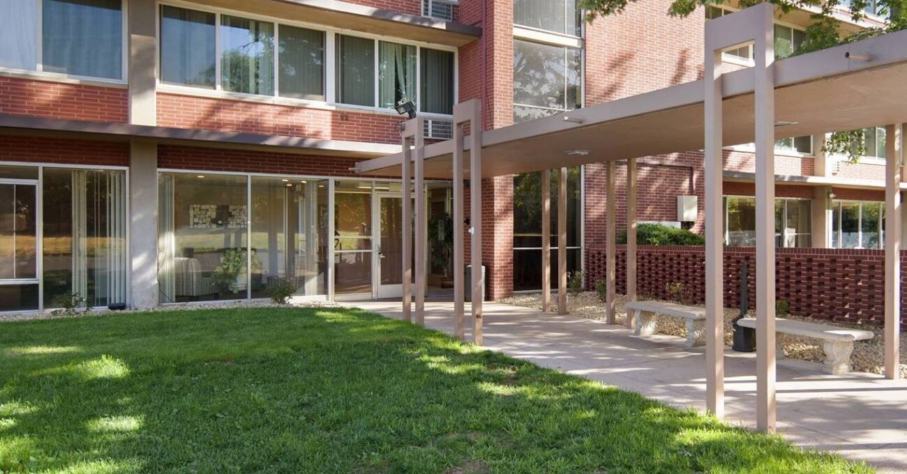 The image shows the entrance of a red-brick building, possibly Columbine Towers, with a covered walkway and green lawn.